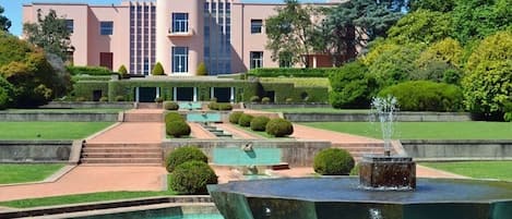 Un edificio moderno con fachada simétrica, una fuente central y un jardín cuidado con setos recortados y un camino despejado hacia la entrada.