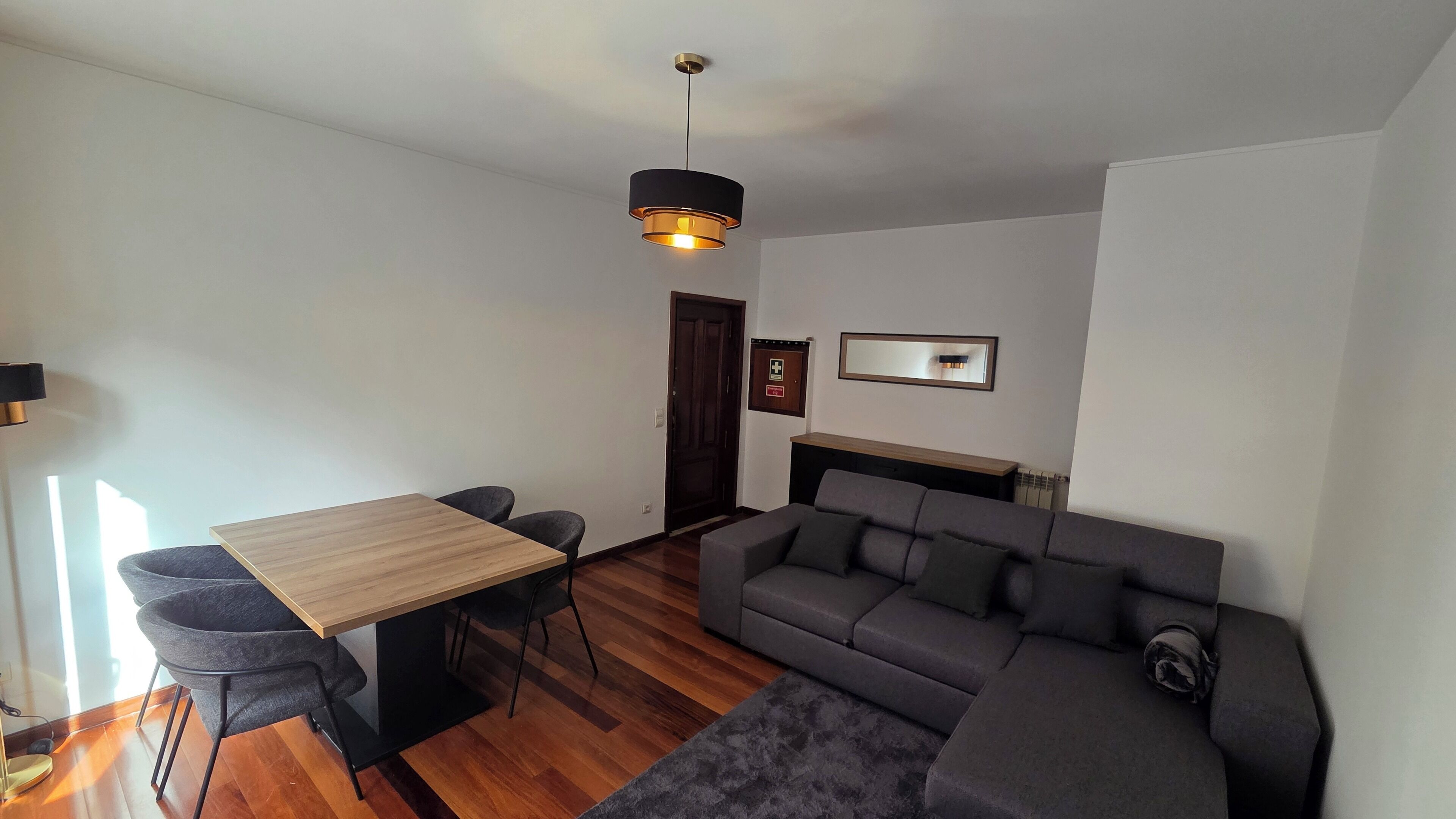 A modern living room with a wooden dining table, two grey armchairs, a grey sofa, and a black pendant light.