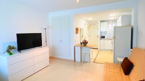 A modern living room with a flat-screen TV, a white entertainment unit, and a sofa with patterned cushions. A kitchen area is visible in the background with white cabinets and a refrigerator. - Colina 10 Studio by Residea (Torremolinos)