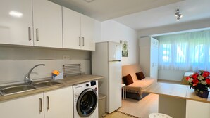 A modern kitchen with a washing machine, a sink, and a refrigerator. There is a small stool and a couch in the living area. - Colina 10 Studio by Residea (Torremolinos)