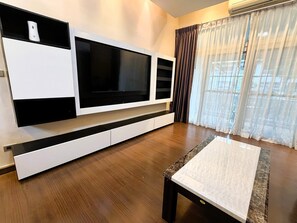 A modern living room with a large flat-screen TV mounted on a white entertainment unit, wooden flooring, and a window with sheer curtains. - Spacious 1-bedroom Condo With Large Balcony at Baan Suan Lalana (Pattaya)
