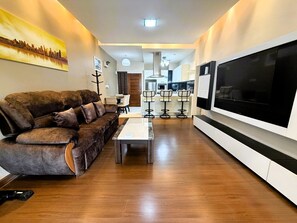 A modern living room with a brown leather sofa, a TV mounted on the wall, and a dining area with bar stools. - Spacious 1-bedroom Condo With Large Balcony at Baan Suan Lalana (Pattaya)