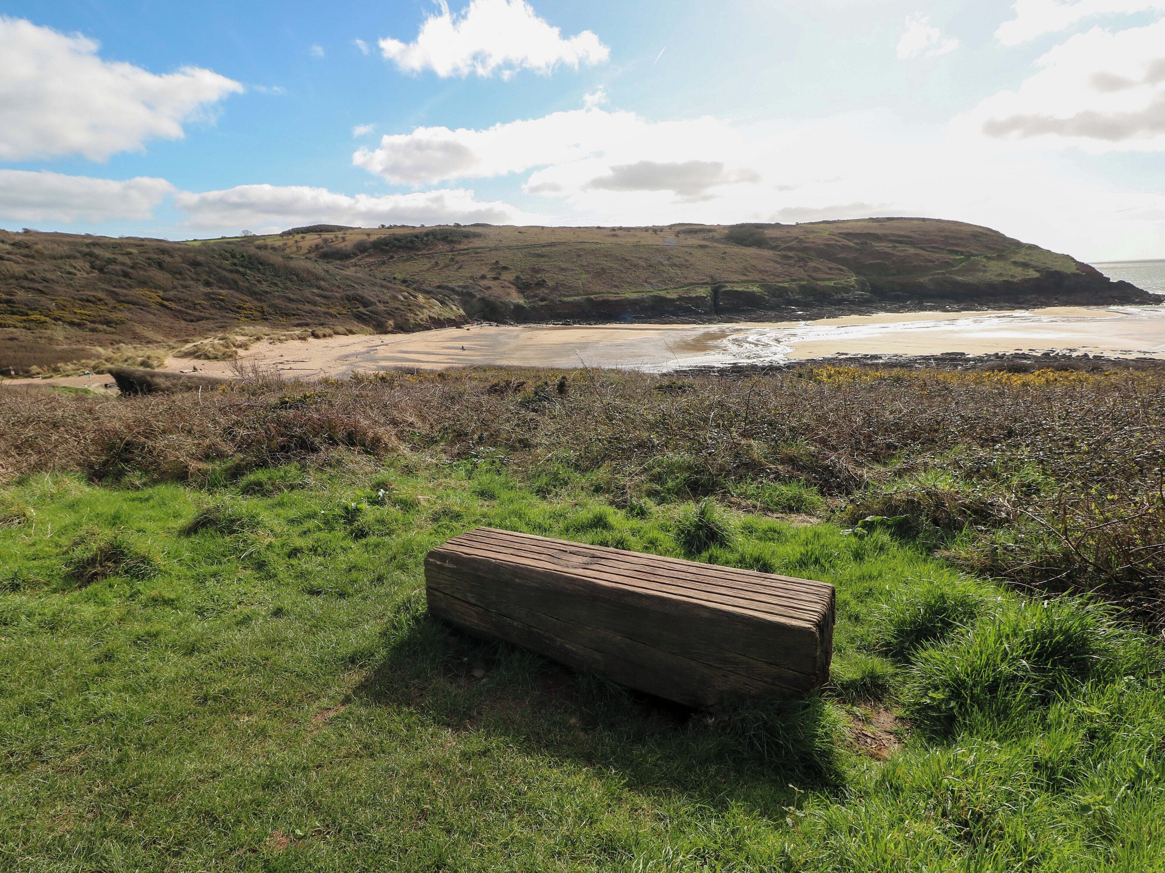 A wooden bench in a grassy area with a view of a beach and hills.