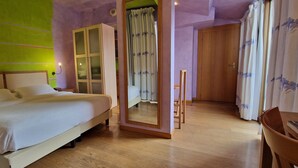 Una cama bien hecha con cabecera, un armario de madera, un espejo y una silla en una habitación con suelo de madera y pared verde.