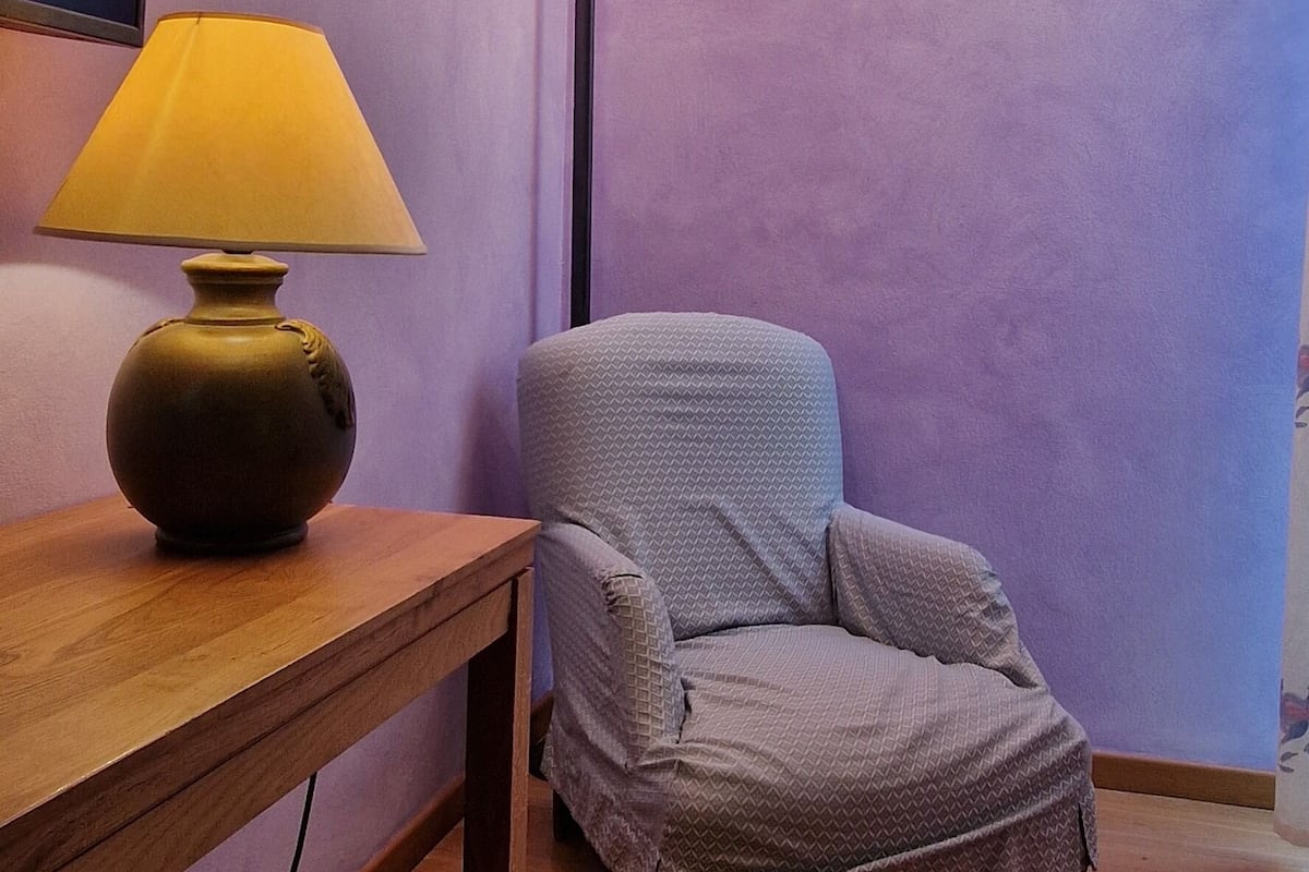 Una habitación con una mesa de madera, una lámpara, un sillón estampado y una luminaria en la pared.