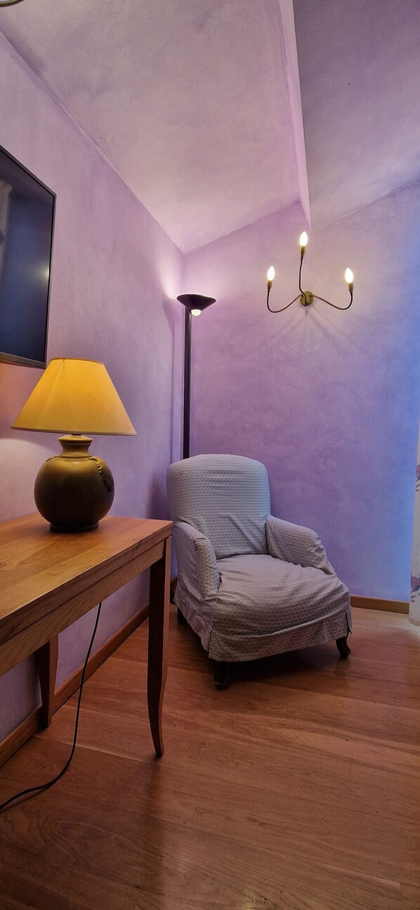 Una habitación con una mesa de madera, una lámpara, un sillón estampado y una luminaria en la pared.