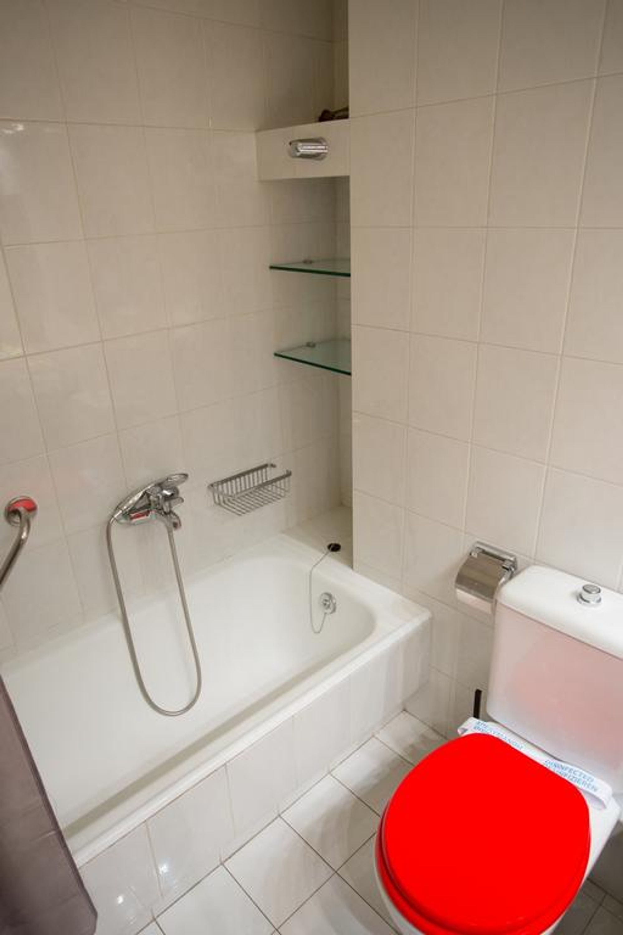 A bathroom with a white bathtub, a red toilet seat, and a glass shelf above the toilet.