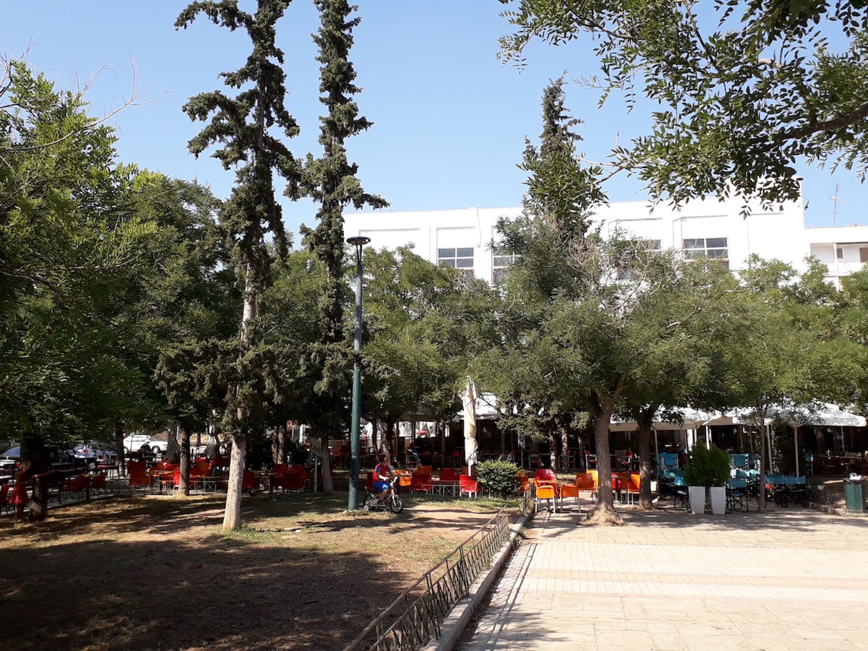 An outdoor seating area with tables and chairs, surrounded by trees and a building in the background.