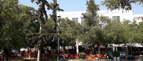 An outdoor seating area with tables and chairs, surrounded by trees and a building in the background.