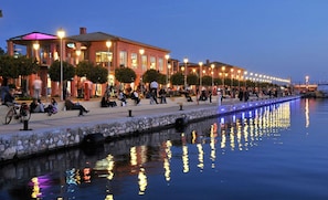 A waterfront promenade with a row of buildings, a canal, and people walking and sitting along the path. - Socrates Path (Athens)