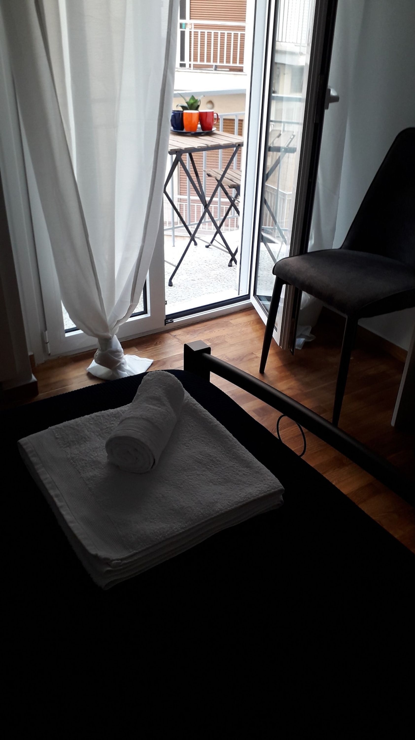 Una stanza con un letto, una sedia, una finestra con tende e vista su un balcone con mobili da esterno.