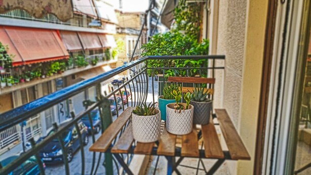 Un balcón con plantas en macetas, una mesa de madera y vista a la calle.
