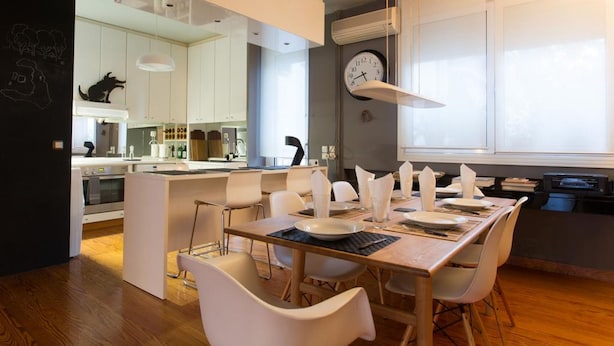 Una cocina moderna con armarios blancos, un comedor con mesa de madera y sillas blancas, y un ventanal con un reloj en la pared.