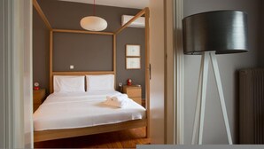 Una cama bien hecha con sábanas blancas y cabecera de madera, una mesita de noche con un reloj rojo y una lámpara de pie moderna.