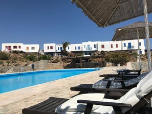 A poolside area with lounge chairs and a clear view of white buildings with blue doors and windows in the background. - Smart 4 Pers Apar in Costa Ilios Resort Myconos (Mykonos)