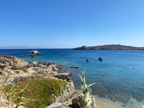 A rocky shoreline with clear blue water, a boat anchored near the shore, and a distant island. - Smart 4 Pers Apar in Costa Ilios Resort Myconos (Mykonos)