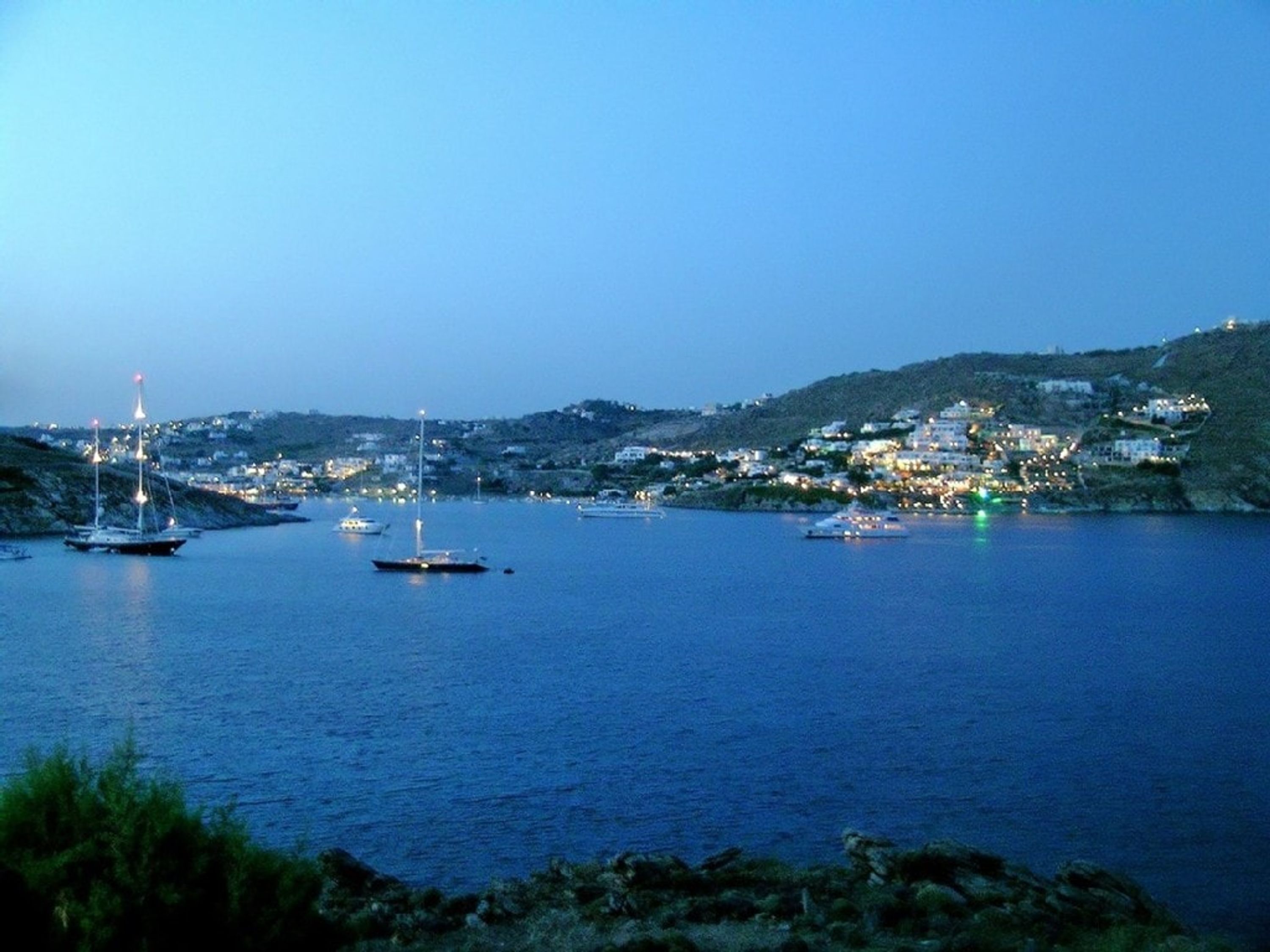 A coastal town at dusk with boats anchored in the harbor and buildings on the hillside.