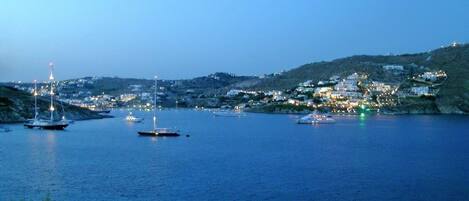 A coastal town at dusk with boats anchored in the harbor and buildings on the hillside.