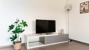 Ein modernes Wohnzimmer mit einem Flachbildfernseher auf einem weißen Schrank, einer Topfpflanze, einer Stehlampe und einem gerahmten Bild an der Wand.