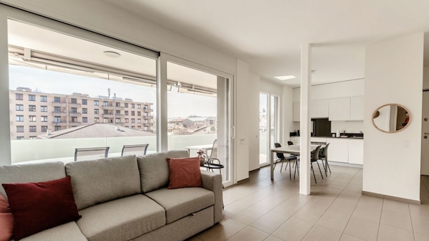 Ein modernes Apartment mit einem großen Schiebeglasfenster, das auf einen Balkon führt, einer Couch mit Kissen, einem Essbereich mit Stühlen und einer Küchenzeile.