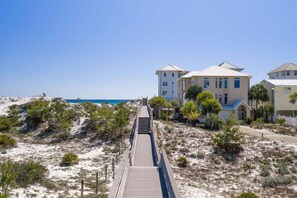 Un hotel frente al mar con un paseo de madera que conduce a la playa, rodeado de dunas de arena y vegetación escasa.