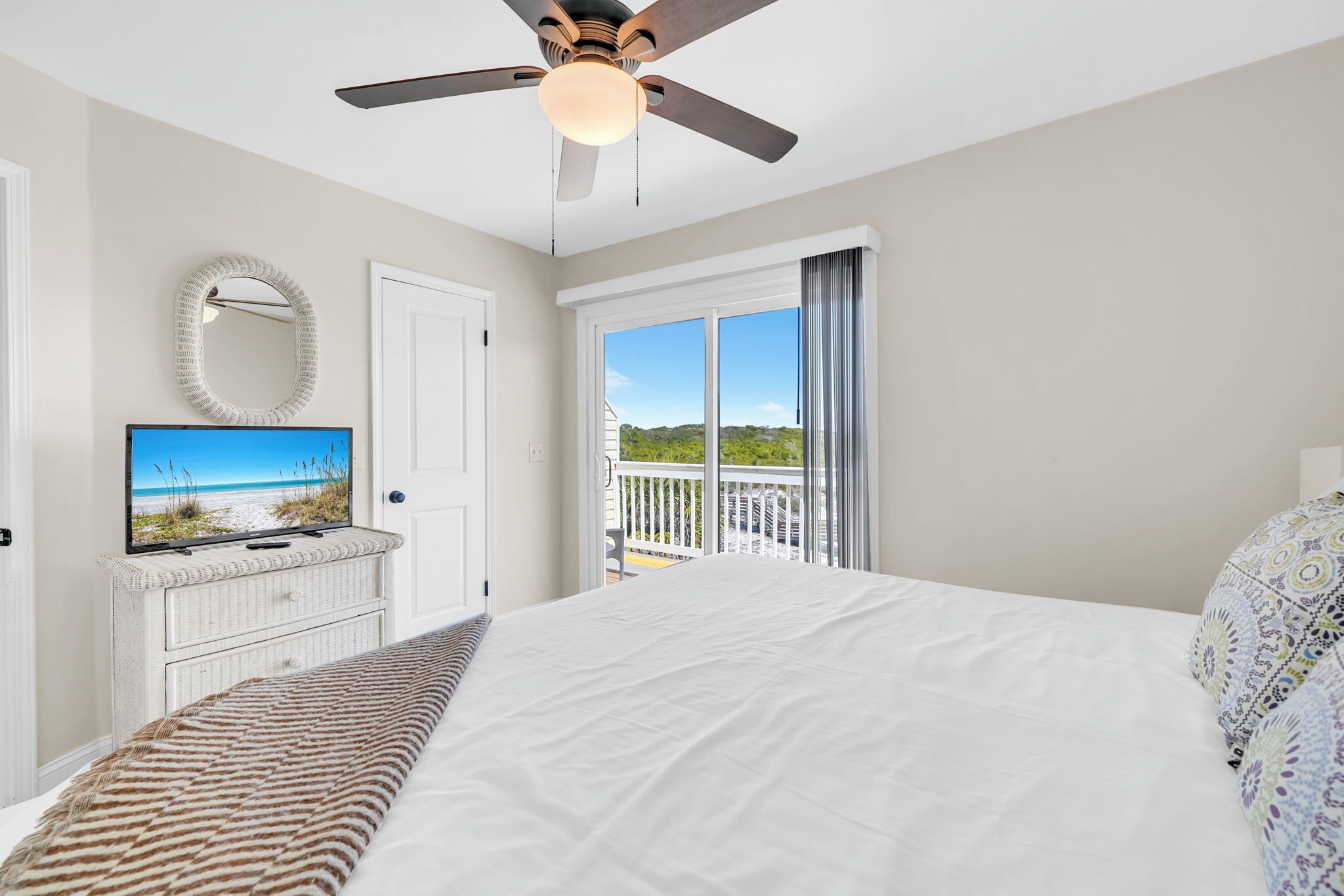 A bedroom with a bed, a ceiling fan, a television on a dresser, a balcony with a view, and a door.