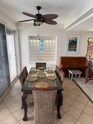 Un coin repas avec une table en verre, des chaises en osier, un ventilateur de plafond, un piano et une vue dégagée sur l’extérieur grâce à une grande fenêtre.