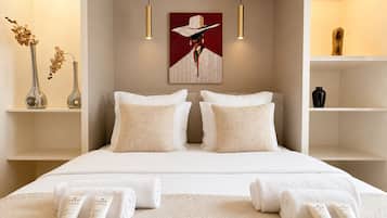 Habitación de hotel con cama, dos mesitas de noche, un cuadro y una estantería con objetos decorativos.