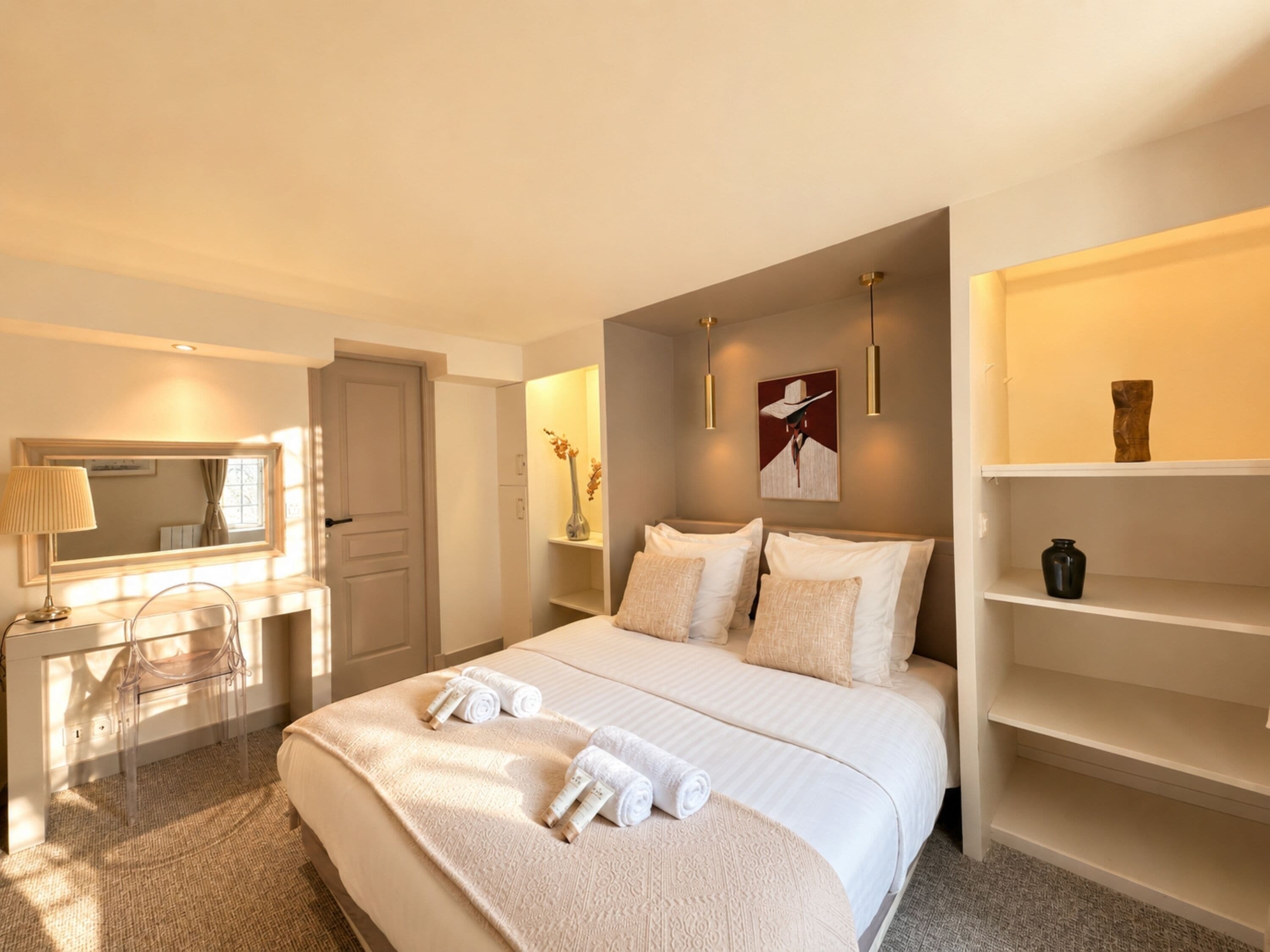 A hotel room with a bed, a desk, a chair, a mirror, and a shelf with decorative items.