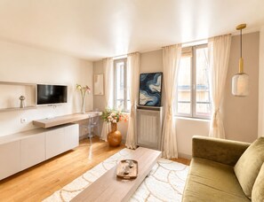 A modern living room with a sofa, a coffee table, a TV, and a dining area with a chair and a vase of flowers. - 1532 Mouffetard (Paris)