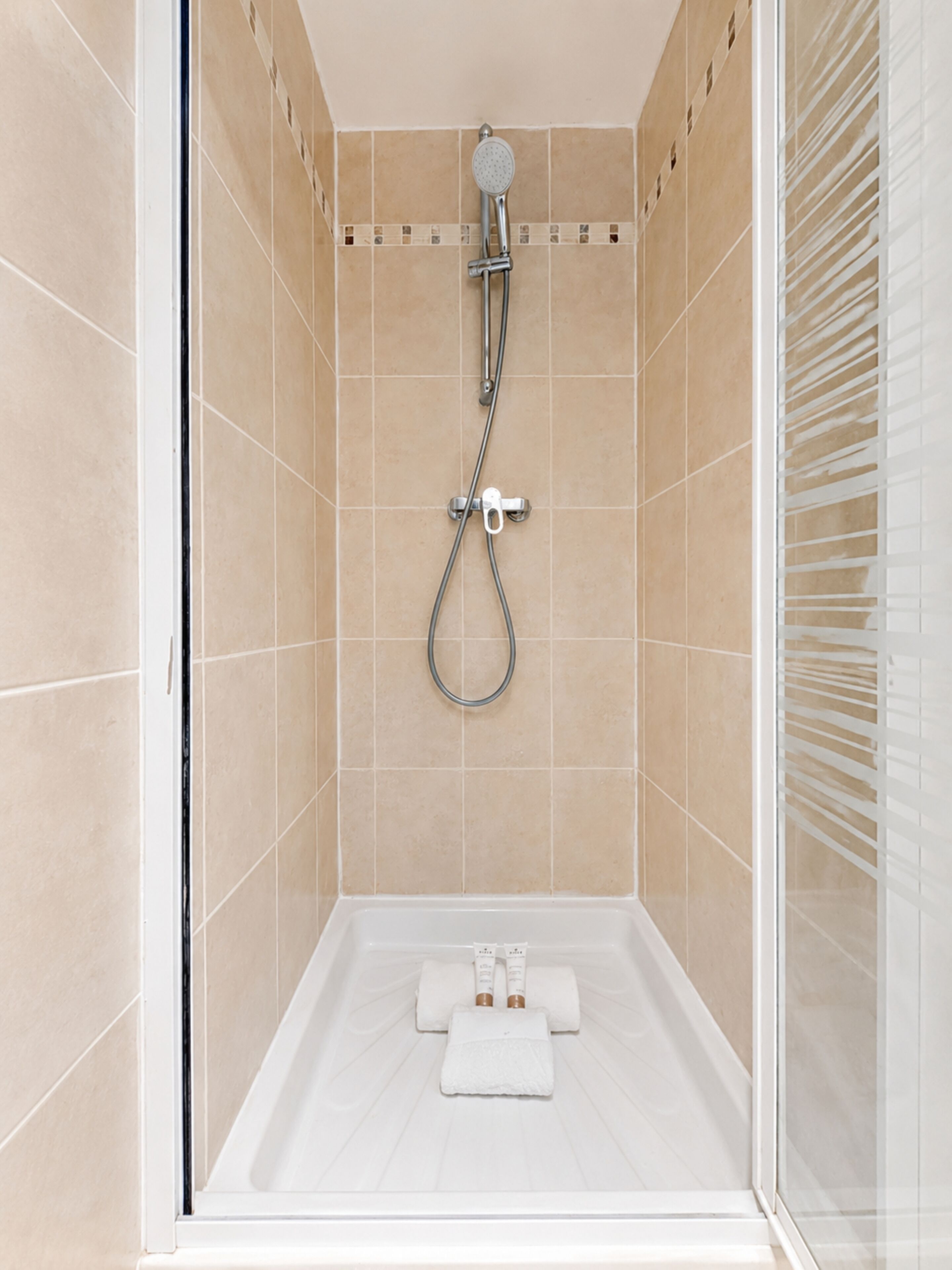 A shower with a handheld showerhead and a wall-mounted faucet, a white bathtub with two towels, and beige tiled walls.