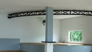 Ein geräumiger Raum mit hoher Decke, einer großen Säule, einem Fenster mit Blick auf Grünflächen und einer schwarzen Metallträgerkonstruktion.