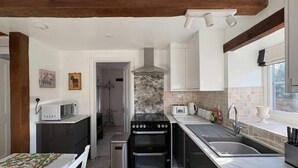 Een kleine keuken met een fornuis, oven, magnetron en spoelbak. Er is een raam dat natuurlijk licht biedt en een deur naar een andere kamer.
