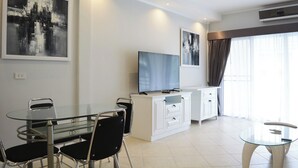 A modern living room with a glass dining table, black chairs, a flat-screen TV, and a white cabinet. - Modern Pool-view 1-bedroom Condo With Large Balcony - Baan Suan Jomtien (Pattaya)