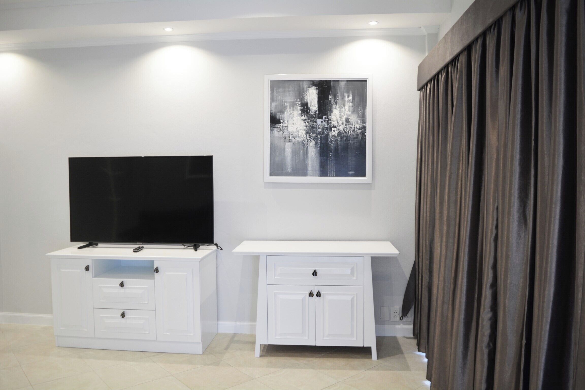 A modern living room with a flat-screen TV on a white cabinet, a framed abstract painting on the wall, and a dark curtain on the right.