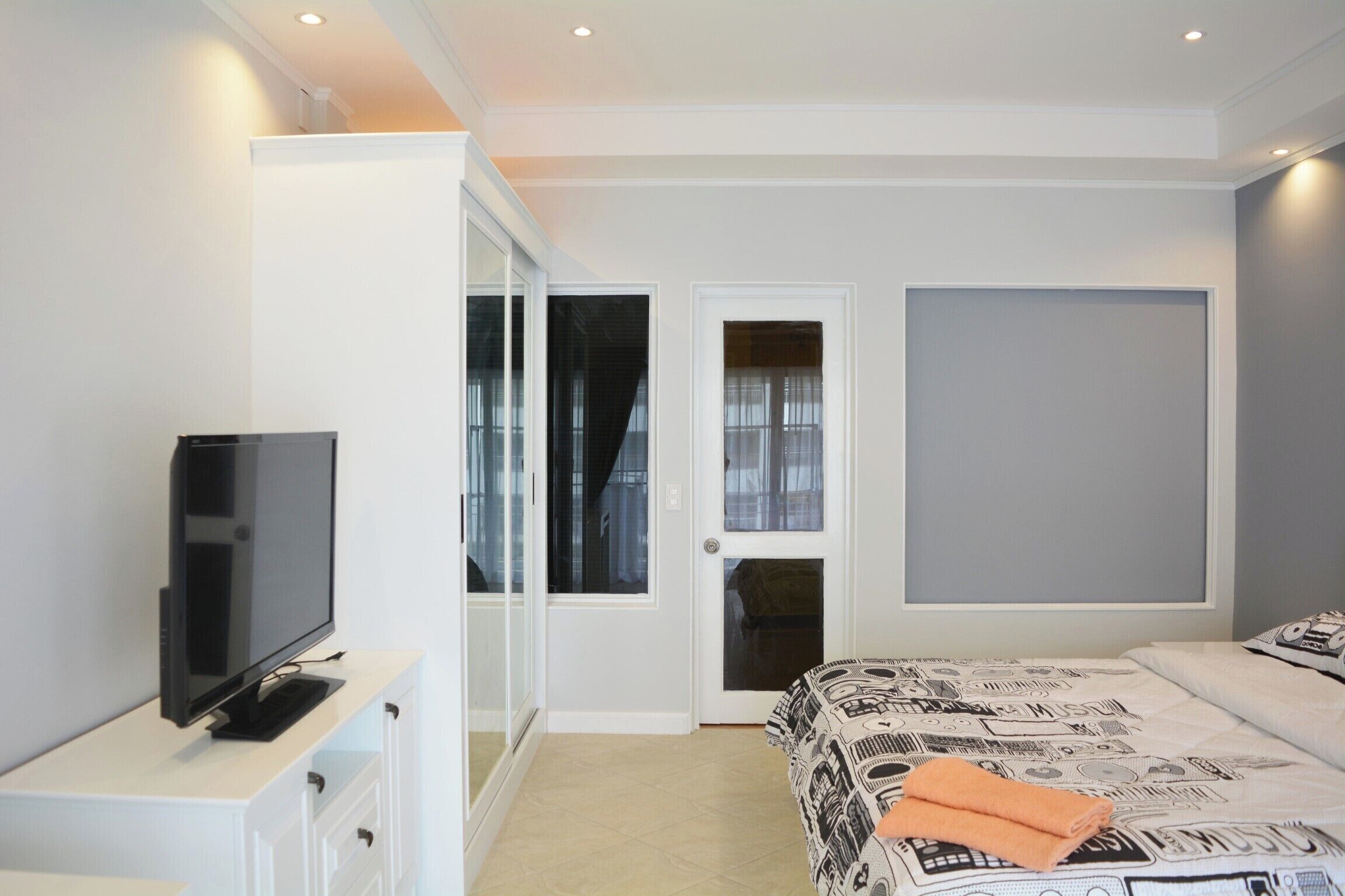 A modern bedroom with a TV on a white cabinet, a bed with patterned bedding, and a door leading to a balcony.