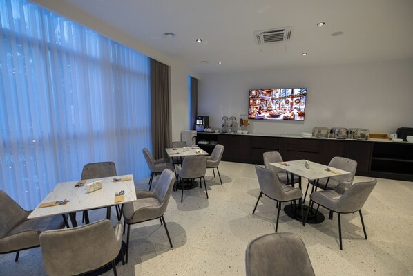 A modern dining area with tables and chairs, a large window with curtains, a flat-screen TV on the wall, and a kitchen area in the background.