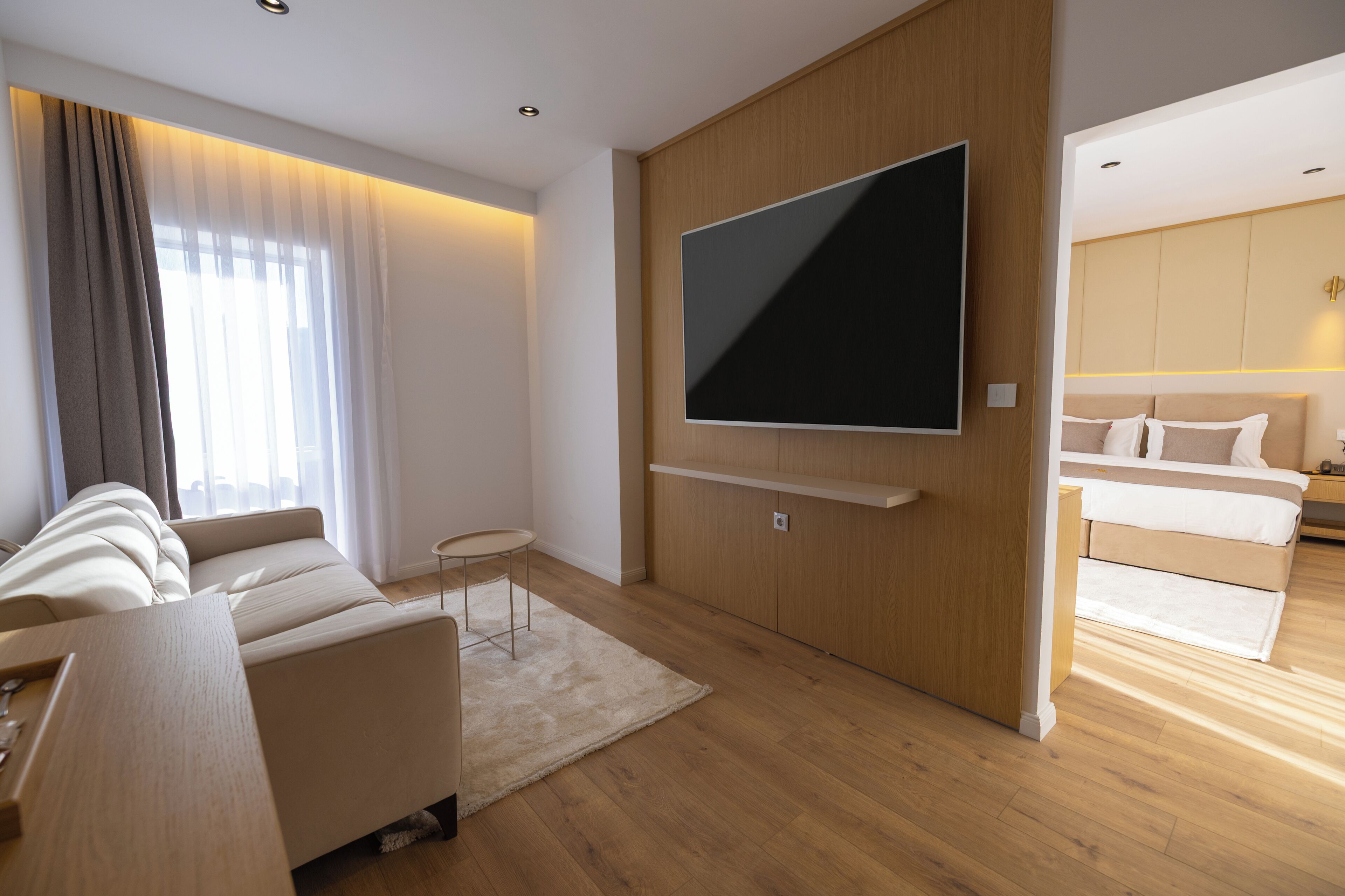 A modern living room with a large flat-screen TV mounted on a wooden wall, a sofa, a small side table, and a view into a bedroom with a bed and nightstands.