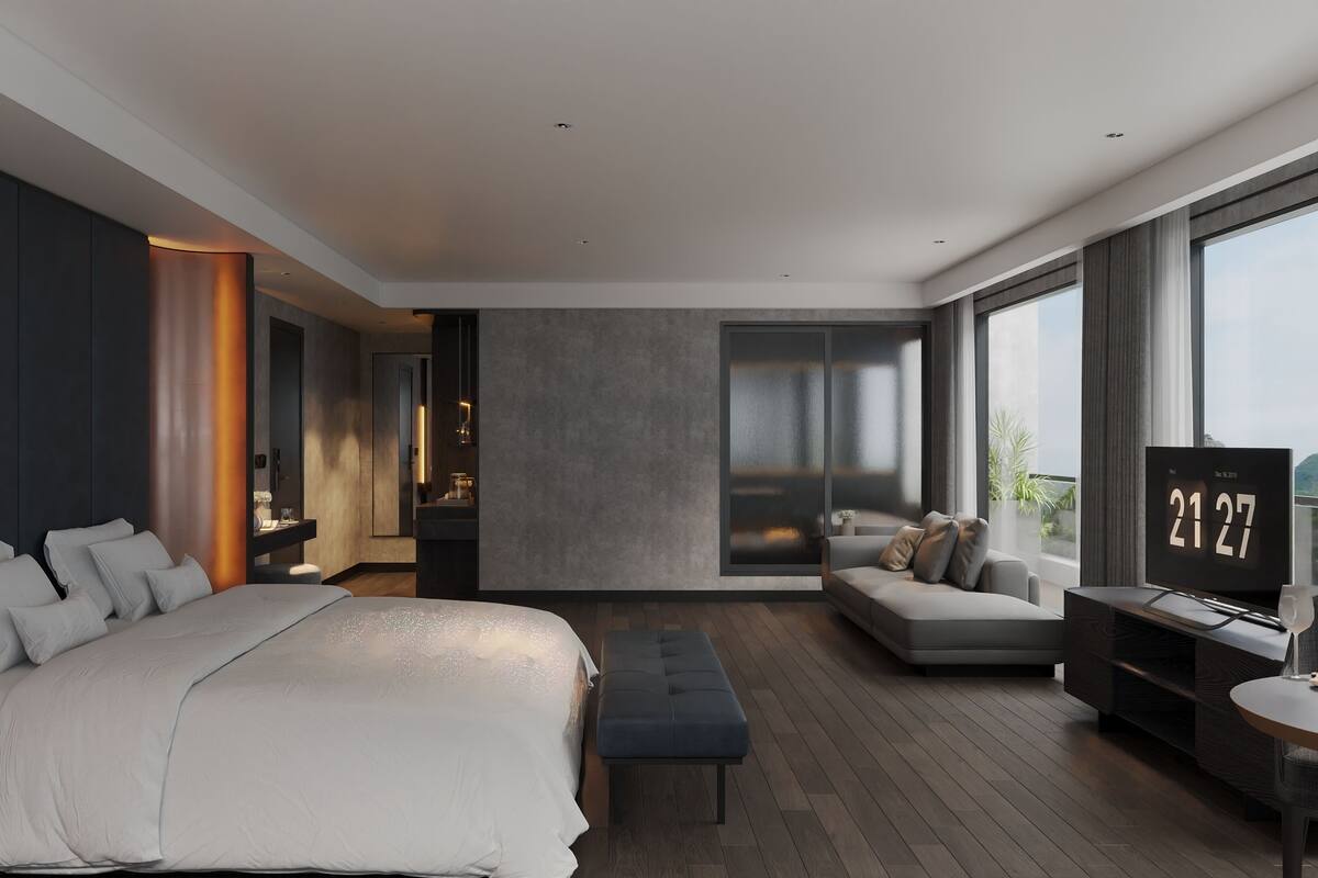 A modern hotel room with a large bed, a seating area with a sofa and armchairs, a small table with a glass, and a TV displaying the date.
