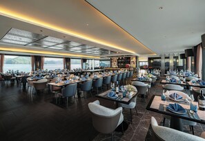 A spacious restaurant with multiple dining tables and chairs, a bar area, and large windows offering a view of the water. - Victorious Cruise Halong Bay (Tuan Chau)