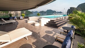 A modern outdoor pool area with lounge chairs, a seating area, and a view of the sea and mountains. - Victorious Cruise (Tuan Chau)