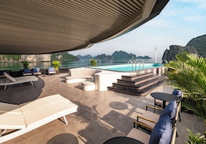 A modern outdoor pool area with lounge chairs, a seating area, and a view of the sea and mountains. - Victorious Cruise (Tuan Chau)