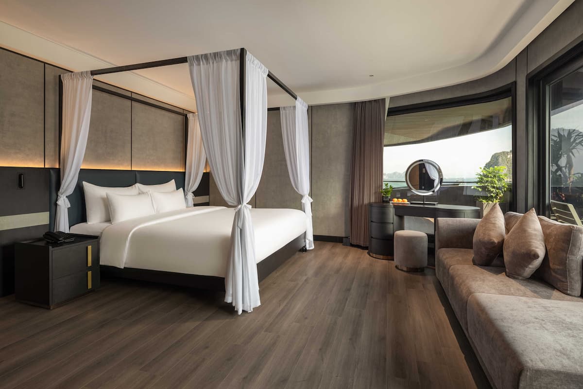 A modern hotel room with a four-poster bed, a sitting area with a sofa, a desk with a mirror, and a view of the outdoors.