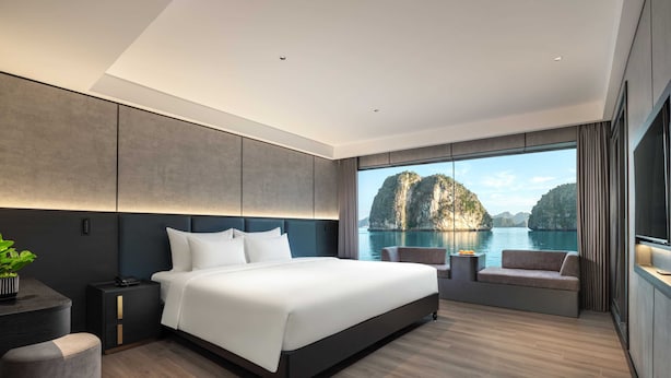A modern bedroom with a large bed, bedside tables, a seating area, and a view of a mountainous landscape. - Victorious Cruise (Tuan Chau)