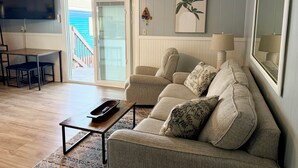 Uma sala de estar com sofá, mesa de centro, televisão e vista através de uma porta de vidro deslizante.