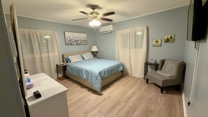 Quarto com cama, uma cadeira, mesa de cabeceira, ventilador de teto e televisão.