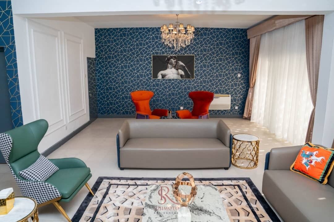 En moderne stue med væg i blå mosaik, en lysekrone, en grå sofa og røde lænestole.