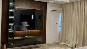 Una camera d'hotel moderna con una grande TV a schermo piatto fissata a una parete rivestita in legno scuro, un letto imbottito rosso, un tappeto decorato e un lampadario.