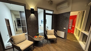 A modern hotel room with a dining area, two chairs, a table with fruit, a sofa, and a large mirror.