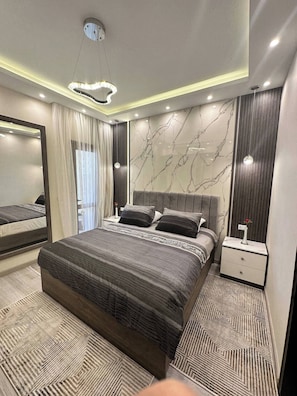 A modern bedroom with a large bed, a nightstand, a mirror, and a marble-patterned wall. - Luxury Apartment- Sahl Hasheesh- Gravity Hotel (Sahl Hasheeh)